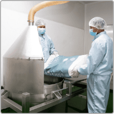 Operators in cleanroom gowning loading raw material into a stainless mixing vessel