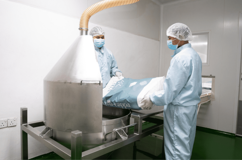 Operators in cleanroom gowning loading raw material into a stainless mixing vessel